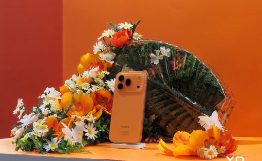 Orange Honor smartphone on a small white stand surrounded by bright orange and white flowers against an orange backdrop.