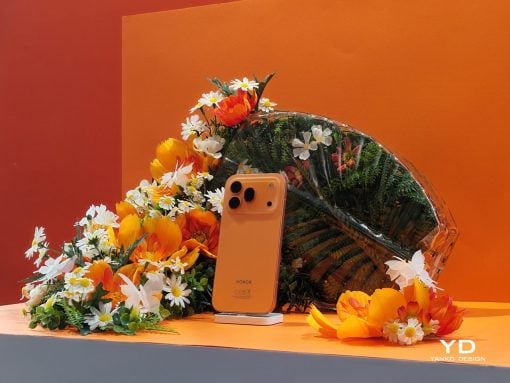 Orange Honor smartphone on a small white stand surrounded by bright orange and white flowers against an orange backdrop.