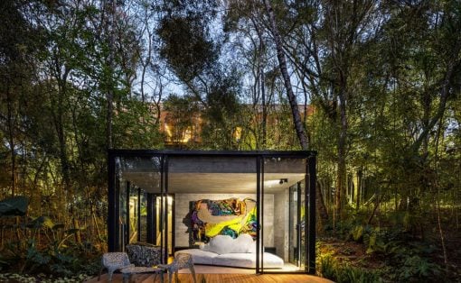 Glass-walled bedroom pavilion in a dense forest, with a white bed and a colorful abstract painting visible inside, on a wooden deck with chairs outside.