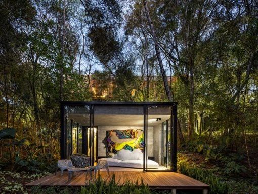 Glass-walled bedroom pavilion in a dense forest, with a white bed and a colorful abstract painting visible inside, on a wooden deck with chairs outside.
