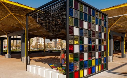 Outdoor pavilion with a colorful glass block wall and yellow supporting beams; a person walks by on a sunny day.