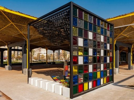 Outdoor pavilion with a colorful glass block wall and yellow supporting beams; a person walks by on a sunny day.