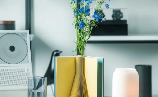 Blue flowers rise from a book-sculpture vase on a white desk, with a glass of water and speaker nearby.