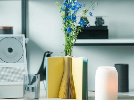 Blue flowers rise from a book-sculpture vase on a white desk, with a glass of water and speaker nearby.