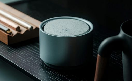 Gray cylindrical smart speaker on a dark wooden desk with blurred countertop items nearby, conveying a modern tech setup.