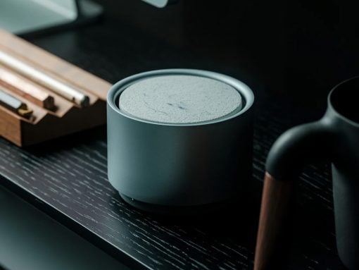 Gray cylindrical smart speaker on a dark wooden desk with blurred countertop items nearby, conveying a modern tech setup.