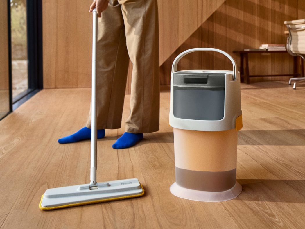 Person mopping a wooden floor with a flat mop, with a beige bucket beside them.