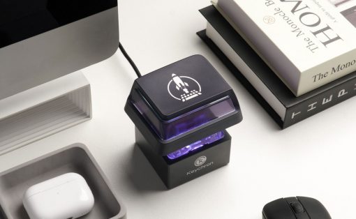 Black self-inking stamp with a purple glow and rocket logo on a white desk beside books and a mouse.