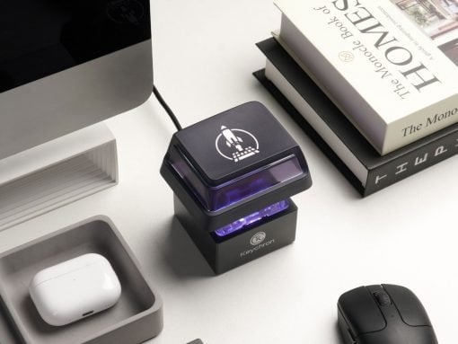 Black self-inking stamp with a purple glow and rocket logo on a white desk beside books and a mouse.