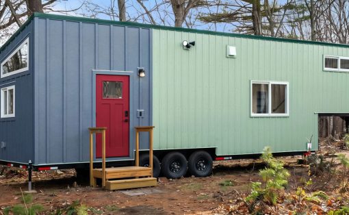 Blue and light-green modular tiny house on a trailer in a forest clearing, red front door with wooden steps outside
