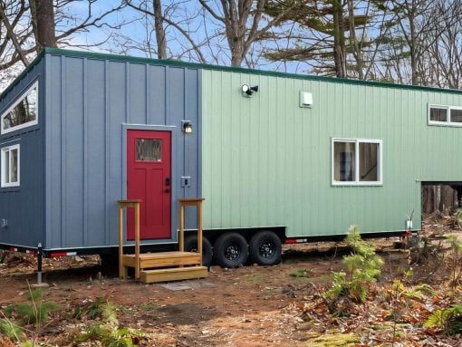 Blue and light-green modular tiny house on a trailer in a forest clearing, red front door with wooden steps outside