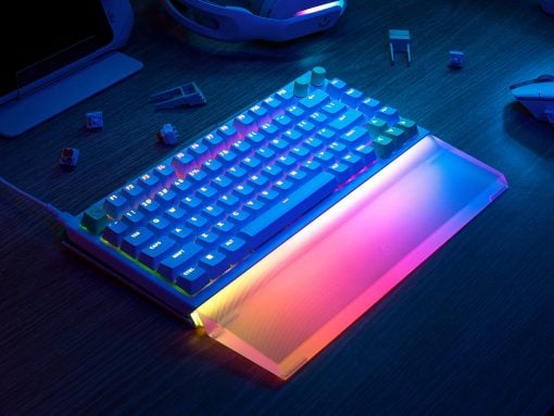 RGB-lit mechanical keyboard with multicolor underglow on a desk, surrounded by peripherals like headphones and a laptop.