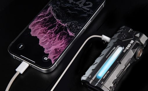 Smartphone connected to a rugged electronic device via a charging cable on a dark surface, blue glow from the device visible.