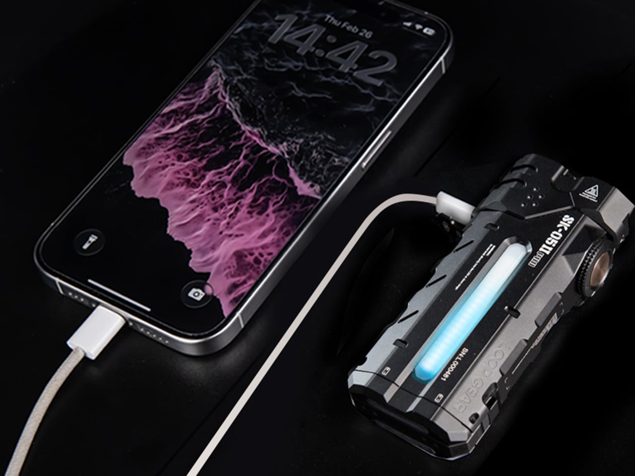 Smartphone connected to a rugged electronic device via a charging cable on a dark surface, blue glow from the device visible.