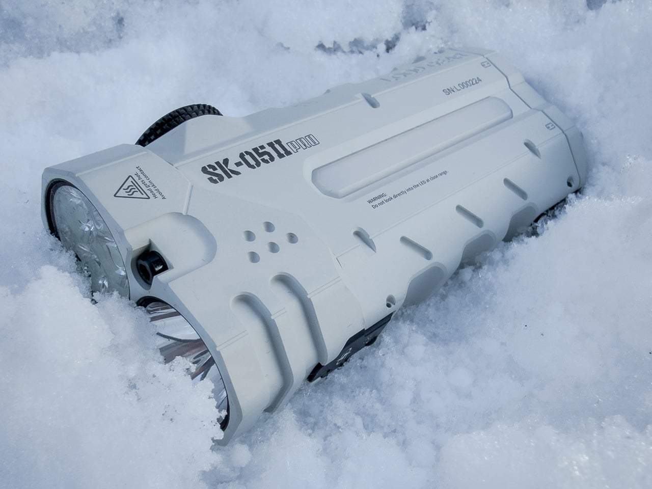 White handheld flashlight partly buried in snow, lens facing left, rugged outdoor design visible.