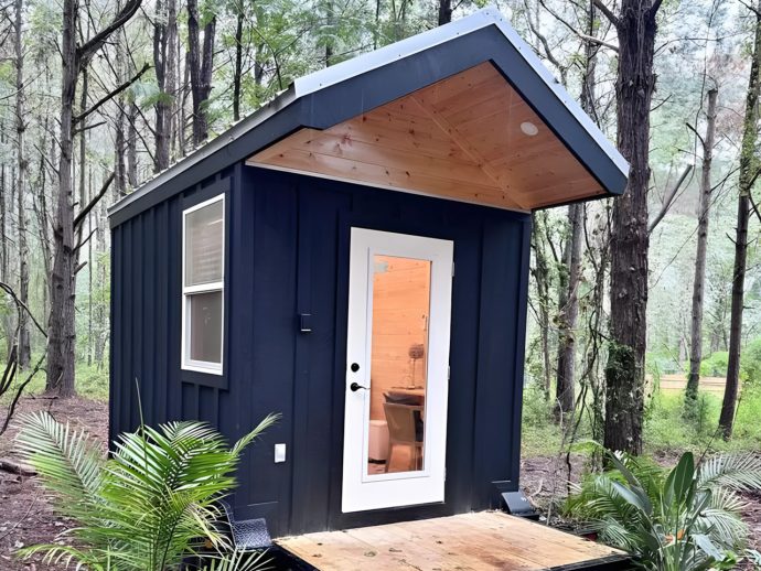Small dark-blue shed in a wooded yard with a white-framed glass door, a wooden deck, and surrounding green plants.