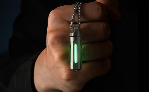 Hand gripping a metallic pendant necklace with a glowing green vertical light inside a small rectangular case against a dark background and chain visible above.