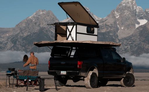 Black pickup with a rooftop camper and pop-up tent parked on a dirt field, two people nearby with camping gear, mountains in the background.