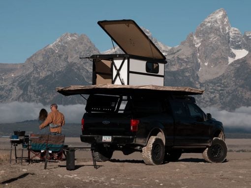 Black pickup with a rooftop camper and pop-up tent parked on a dirt field, two people nearby with camping gear, mountains in the background.