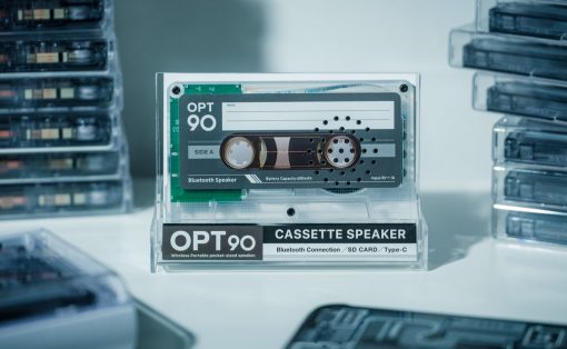 Clear display case housing an OPT90 cassette speaker with Bluetooth label and cassette graphic, stacks of tapes in the background.