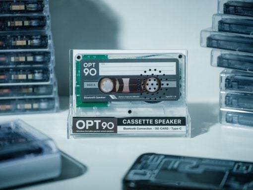 Clear display case housing an OPT90 cassette speaker with Bluetooth label and cassette graphic, stacks of tapes in the background.