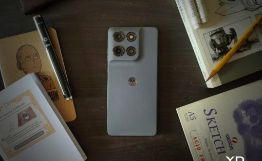 Gray Motorola smartphone with a quad-camera back sits on a dark wooden desk, surrounded by notebooks, sketches, and a pencil.