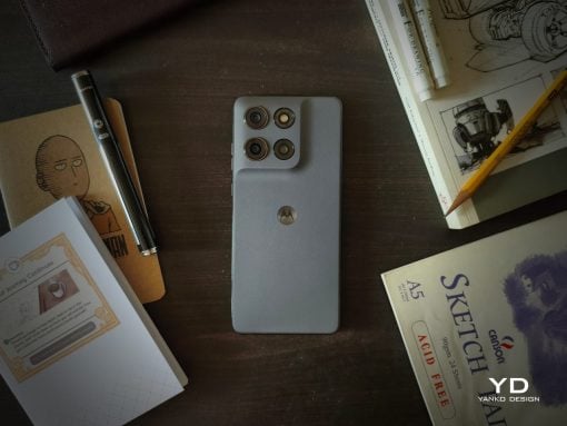 Gray Motorola smartphone with a quad-camera back sits on a dark wooden desk, surrounded by notebooks, sketches, and a pencil.