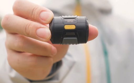 Close-up of a hand holding a small black wearable device with a yellow button on top.