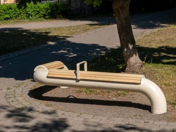 Curved white pipe bench with wooden slats beside a tree in a park setting