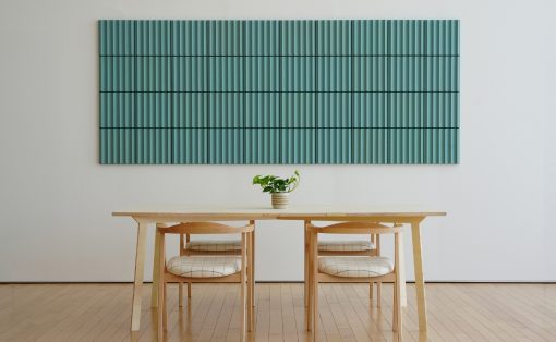 Minimalist dining area with a light wood table and two chairs, teal vertical-slatted wall art, and a small plant centerpiece.