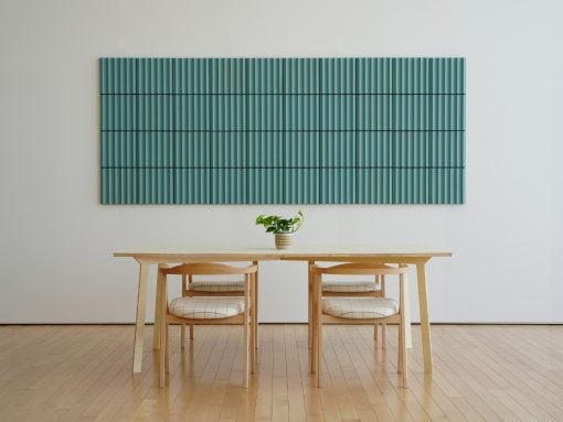 Minimalist dining area with a light wood table and two chairs, teal vertical-slatted wall art, and a small plant centerpiece.