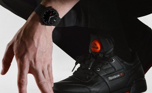 Close-up of a black Reebok sneaker with orange tongue and laces, worn with a black outfit; a wristwatch is nearby.