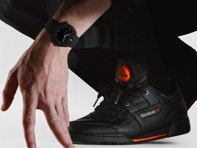 Close-up of a black Reebok sneaker with orange tongue and laces, worn with a black outfit; a wristwatch is nearby.