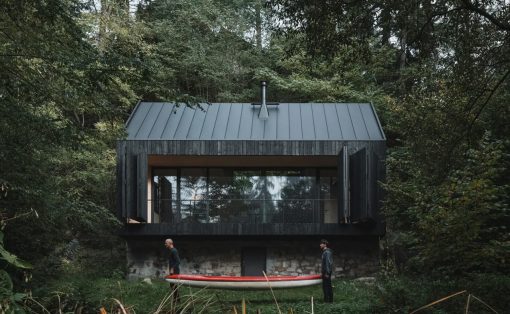 Modern black wooden cabin on a stone foundation nestled in a dense forest, with two people standing by a red-and-white canoe outside.