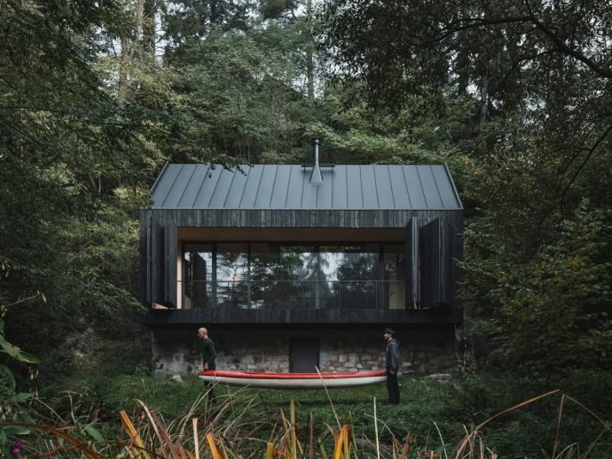 Modern black wooden cabin on a stone foundation nestled in a dense forest, with two people standing by a red-and-white canoe outside.