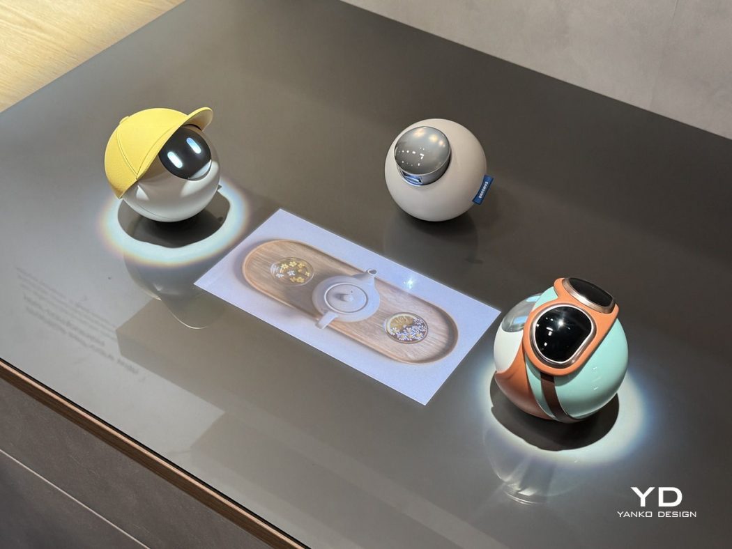 Three spherical design robots on a display table: yellow-cap orb, gray orb with blue tag, and mint-orange teal orb with a dark lens, centered around a printed design card. The Yanko Design logo is in the bottom right.