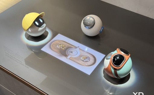 Three spherical design robots on a display table: yellow-cap orb, gray orb with blue tag, and mint-orange teal orb with a dark lens, centered around a printed design card. The Yanko Design logo is in the bottom right.