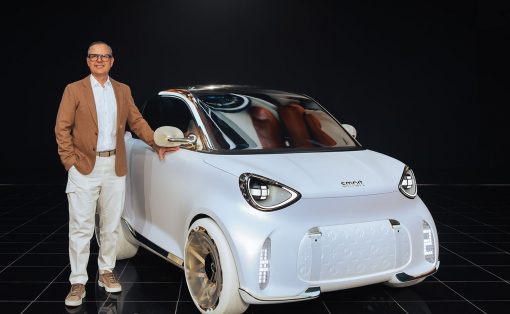 Man in a tan blazer and white pants stands beside a small futuristic white Smart car in a dark studio backdrop.