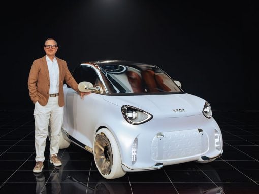 Man in a tan blazer and white pants stands beside a small futuristic white Smart car in a dark studio backdrop.