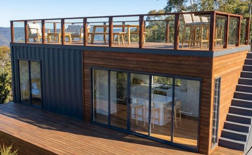 Modern two-story house with dark blue metal siding and wood-clad exterior, large sliding glass doors, and a rooftop deck with chairs and a glass railing.