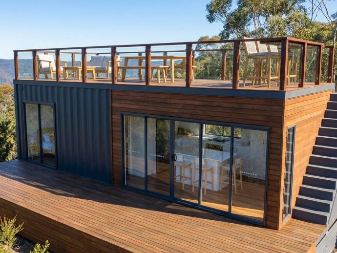 Modern two-story house with dark blue metal siding and wood-clad exterior, large sliding glass doors, and a rooftop deck with chairs and a glass railing.