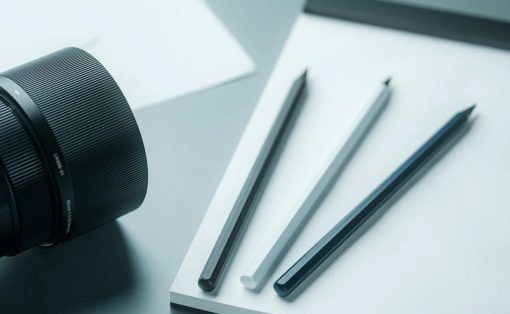 Close-up of a camera lens beside a white notebook with three pencils on the page surface.
