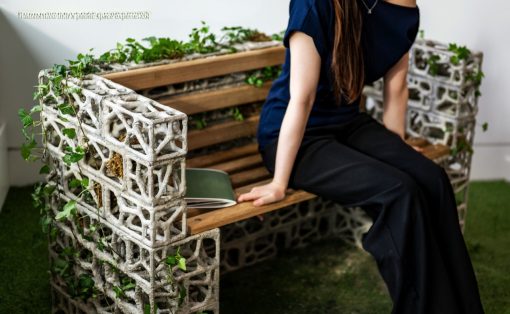 Woman sits on an industrial-style bench made from concrete blocks and wooden slats, ivy winding along the sides, in a bright indoor space with green carpet.