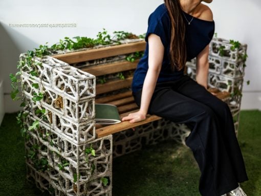 Woman sits on an industrial-style bench made from concrete blocks and wooden slats, ivy winding along the sides, in a bright indoor space with green carpet.