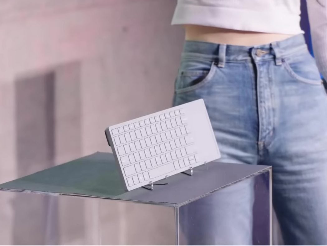 Tiny white keyboard displayed on a metal stand atop a reflective table, with a person in jeans in the background.
