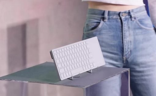 Tiny white keyboard displayed on a metal stand atop a reflective table, with a person in jeans in the background.