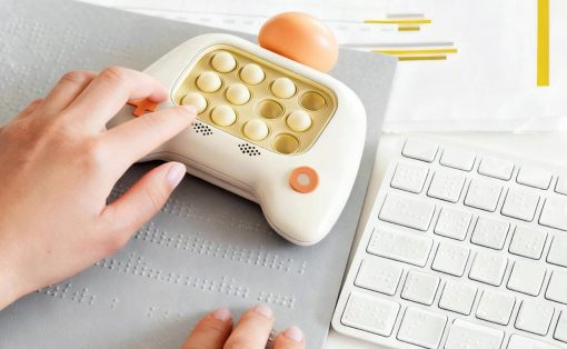 Hand placing an orange insert into a white pill organizer with round compartments on a desk beside a white keyboard and braille mat.