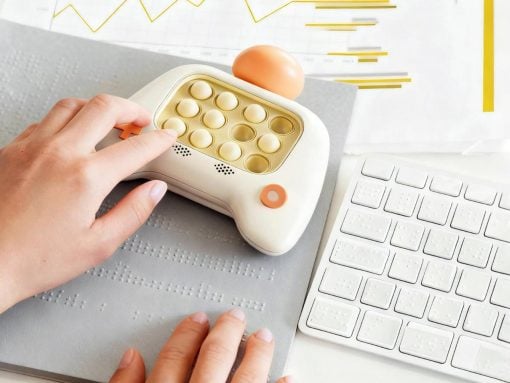 Hand placing an orange insert into a white pill organizer with round compartments on a desk beside a white keyboard and braille mat.