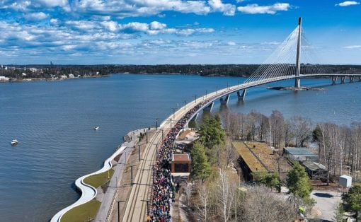 Crowd of people walking along a curved waterfront bridge over a wide river, with a cable-stayed bridge in the background under a blue sky.