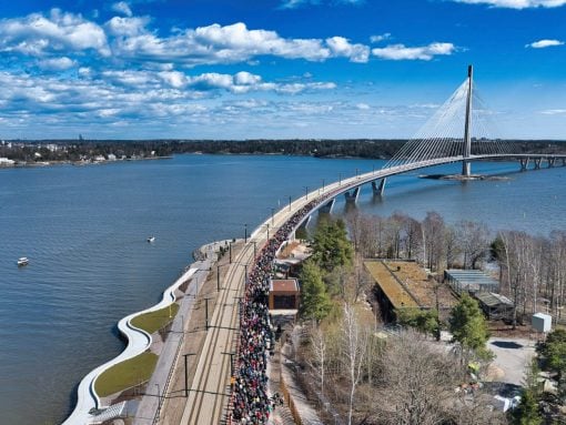 Crowd of people walking along a curved waterfront bridge over a wide river, with a cable-stayed bridge in the background under a blue sky.